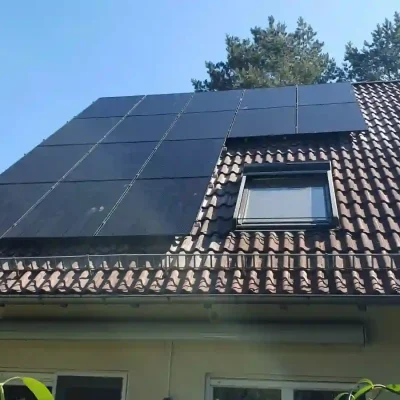 Solarpanels neben einem Dachfenster auf einem braunen Ziegeldach, von Bäumen im Hintergrund umrahmt. Komplette Photovoltaikanlage inkl. Montage und Anmeldung Solarfirma in der nähe