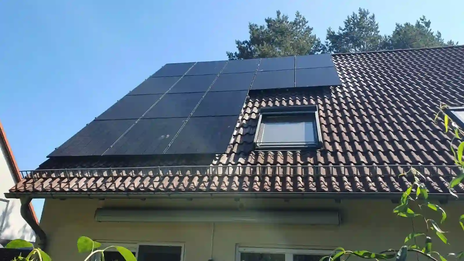Solarpanels neben einem Dachfenster auf einem braunen Ziegeldach, von Bäumen im Hintergrund umrahmt. Komplette Photovoltaikanlage inkl. Montage und Anmeldung