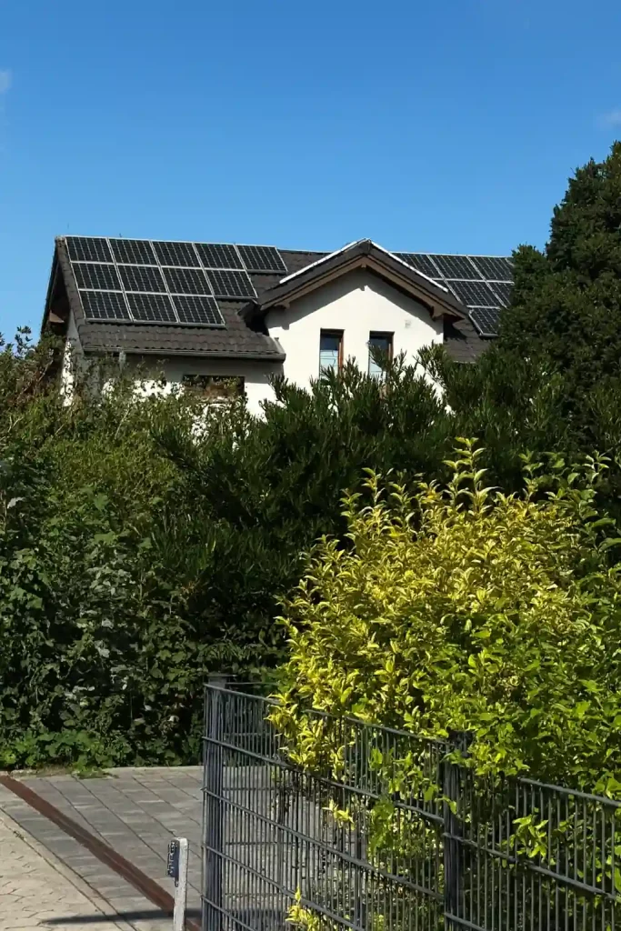 Auf dem Bild ist ein Einfamilienhaus zu sehen, dessen Dachflächen mit Solarmodulen bestückt sind. Die dunklen Photovoltaikmodule sind gleichmäßig auf dem Satteldach angeordnet und wirken modern und unauffällig. Um das Haus herum befinden sich dichte grüne Hecken und Sträucher, die einen natürlichen Rahmen bilden. Der Himmel ist klar und blau – ideales Wetter für Solarstrom. Die Szene zeigt eindrucksvoll, wie eine kleine Photovoltaikanlage in ein typisches Wohnumfeld integriert werden kann: Sie nimmt wenig Platz ein, fügt sich harmonisch in die Dachlandschaft ein und liefert gleichzeitig sauberen, nachhaltigen Strom für den Haushalt.