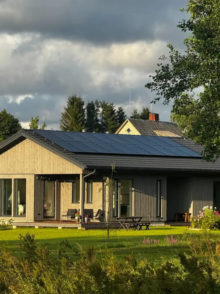 Das Bild zeigt ein modernes Einfamilienhaus mit Holzfassade und großen Glasfronten. Auf dem Dach ist eine Solaranlage für Einfamilienhaus - Einfamilienhaus Solaranlage installiert, die sich harmonisch in die Architektur einfügt. Das Haus steht in einer grünen Umgebung mit gepflegtem Rasen, Blumen und Bäumen. Im Vordergrund erkennt man eine Terrasse mit Sitzmöbeln, die zum Verweilen einlädt. Im Hintergrund sorgen Bäume und ein teils bewölkter Himmel für eine natürliche und stimmungsvolle Atmosphäre. PV Anlage planen