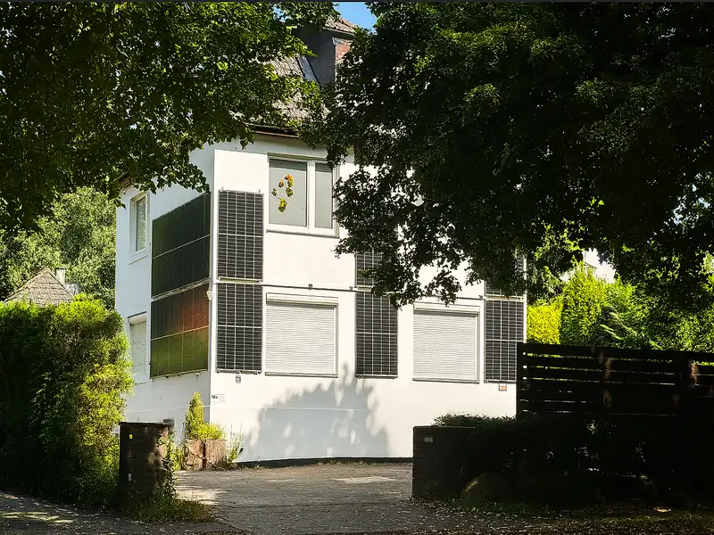 Das Foto zeigt ein modernes, zweistöckiges weißes Wohnhaus mit an der Fassade angebrachten Solarpanelen. Die Szene ist bei Tageslicht aufgenommen und von üppigem Grün umrahmt. Im Vordergrund verläuft eine gepflasterte Einfahrt, die harmonisch in die Umgebung übergeht. Das zuvor sichtbare Fahrzeug wurde entfernt, sodass der Blick frei auf die architektonischen Details des Hauses sowie die gepflegte, naturnahe Umgebung fällt. Blumenverzierte Fensterläden und eine kleine Holzeinzäunung verleihen dem Bild eine wohnliche Atmosphäre. Fassaden PV Anlage mit Montage und Anmeldung.