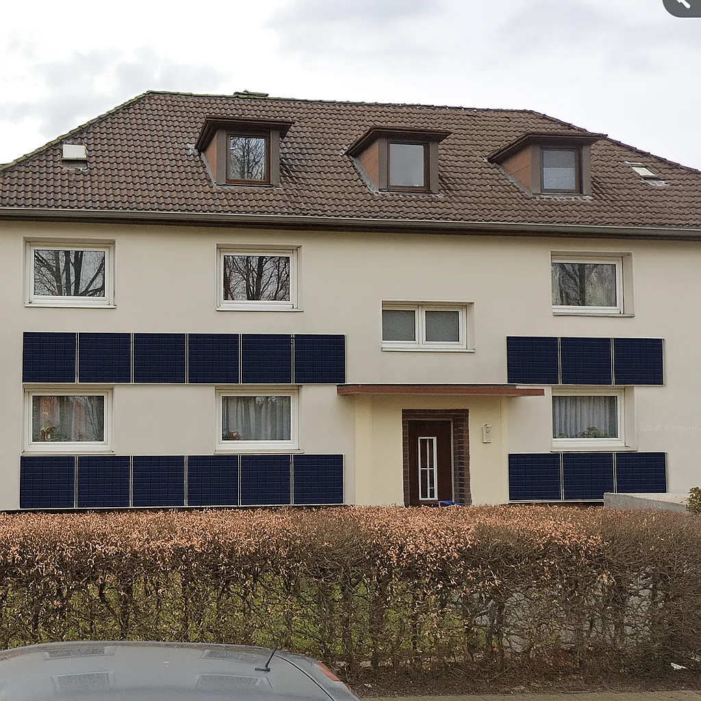 Das Bild zeigt ein Mehrfamilienhaus mit Satteldach, an dessen Fassade eine Fassaden-PV-Anlage mit Montage und Anmeldung installiert wurde. Die Photovoltaikmodule sind in mehreren Reihen horizontal an der Vorderseite des Gebäudes angebracht – sowohl im Erdgeschoss als auch im ersten Obergeschoss. Die dunkelblauen Solarpaneele kontrastieren deutlich mit der hellen Putzfassade und sind unterhalb sowie zwischen den Fenstern angebracht. Die Anlage wirkt professionell montiert und ist optisch gut in die Architektur integriert. Der Eingangsbereich befindet sich mittig unter einem kleinen Vordach. Das Dachgeschoss verfügt über drei Gaubenfenster. Im Vordergrund ist eine gepflegte Hecke zu sehen, die das Grundstück zur Straße hin abgrenzt. Das Bild vermittelt einen Eindruck von moderner, nachhaltiger Energiegewinnung direkt an der Gebäudehülle.