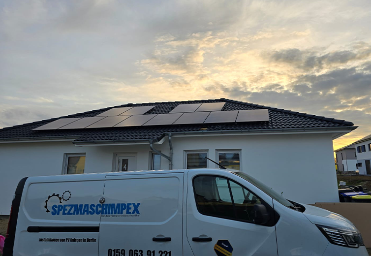 Das Bild zeigt ein modernes Einfamilienhaus mit installierter Photovoltaikanlage auf dem Dach. Die PV-Module sind sauber und flächenbündig in mehreren Reihen auf dem dunklen Walmdach montiert und wirken fachgerecht installiert. Im Vordergrund steht ein weißer Firmen-Transporter mit dem Logo „SPEZMASCHIMPEX“. Auf dem Fahrzeug ist außerdem der Hinweis „Installationen von PV Anlagen in Berlin“ sowie eine Telefonnummer zu sehen. Der Transporter unterstreicht den professionellen Montage- und Installationsbezug. Das Haus befindet sich offenbar in einem Neubaugebiet; rechts sind weitere moderne Wohnhäuser erkennbar. Der Himmel ist teilweise bewölkt und durch warmes Licht geprägt, was auf eine Aufnahme in den Abend- oder frühen Morgenstunden hindeutet. Insgesamt vermittelt das Bild einen professionellen Eindruck einer fertiggestellten PV-Installation im Wohnbereich und eignet sich gut für Referenz- oder Marketingzwecke.