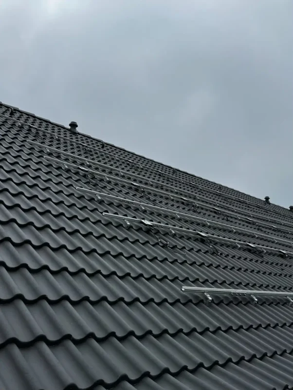 Das Bild zeigt ein Schrägdach mit dunkelgrauen Dachziegeln unter einem bewölkten Himmel. Auf der Dachfläche sind bereits silberfarbene Metallschienen montiert, die parallel zueinander verlaufen. Dabei handelt es sich um die Unterkonstruktion für eine PV-Anlage, auf der später Solarmodule befestigt werden.