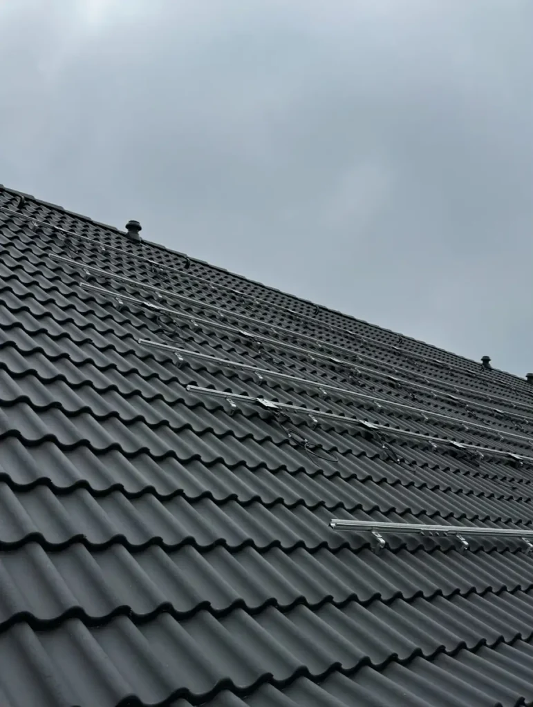 Das Bild zeigt ein Schrägdach mit dunkelgrauen Dachziegeln unter einem bewölkten Himmel. Auf der Dachfläche sind bereits silberfarbene Metallschienen montiert, die parallel zueinander verlaufen. Dabei handelt es sich um die Unterkonstruktion für eine PV-Anlage, auf der später Solarmodule befestigt werden.